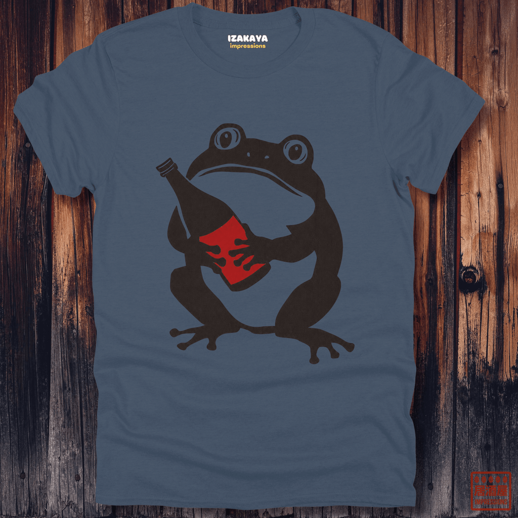 A slate blue crewneck t-shirt featuring a graphic of a dark brown frog holding a red bottle, displayed flat against a rustic dark wood plank background. The brand name 'IZAKAYA' is printed inside the collar.